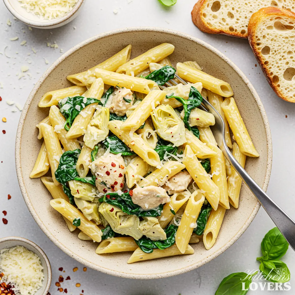 An overhead shot of a rustic bowl filled with creamy chicken, spinach, and artichoke penne pasta, garnished with Parmesan cheese and red pepper flakes, with slices of bread on the side.