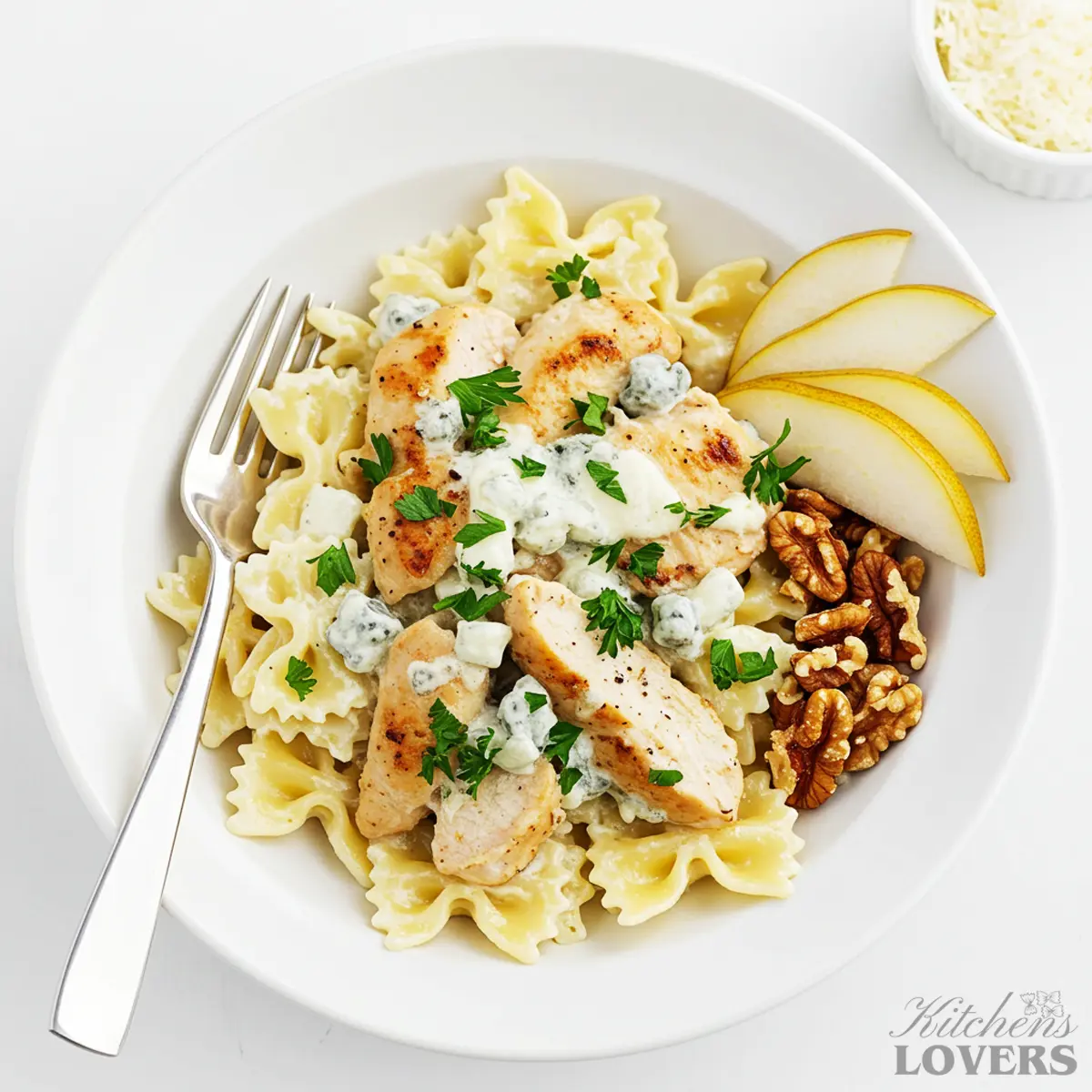 A white bowl filled with creamy chicken gorgonzola pasta with farfalle noodles. The dish is garnished with fresh parsley and served with fresh pear slices and walnuts on the side.