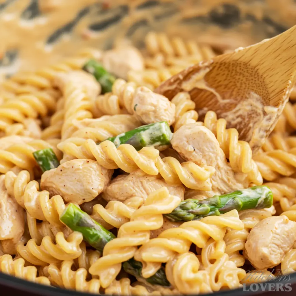 A close-up action shot of a wooden spoon mixing creamy chipotle pasta with chicken and asparagus in a skillet.