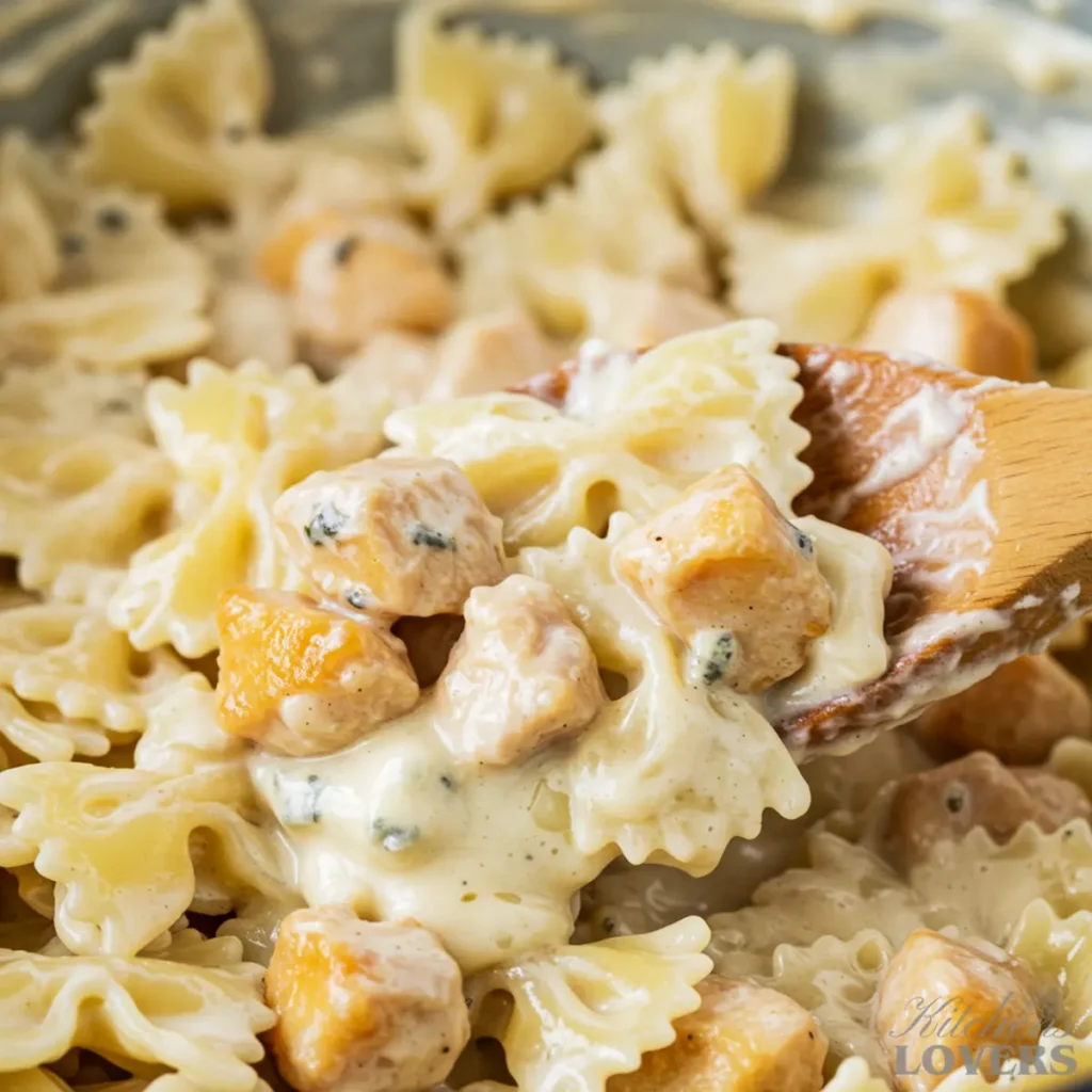 A detailed macro photo showing a wooden spoon stirring farfalle pasta and chicken in a rich, creamy gorgonzola sauce in a skillet.