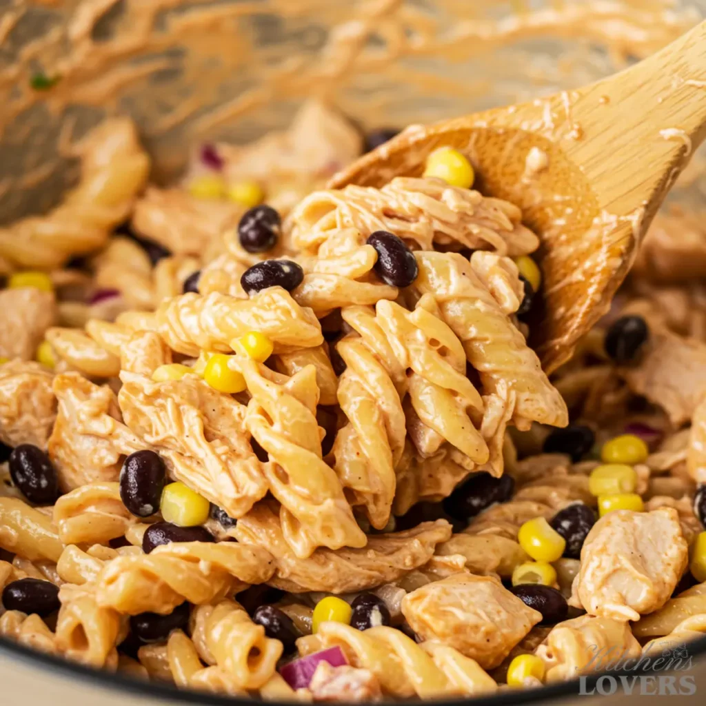 A close-up action shot of pasta, chicken, corn, and beans being mixed with a wooden spoon, showing the creamy texture of the dressing.