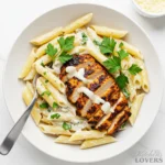 An overhead shot of a white bowl filled with creamy blackened chicken pasta, topped with sliced chicken, fresh parsley, and a side of Parmesan cheese.