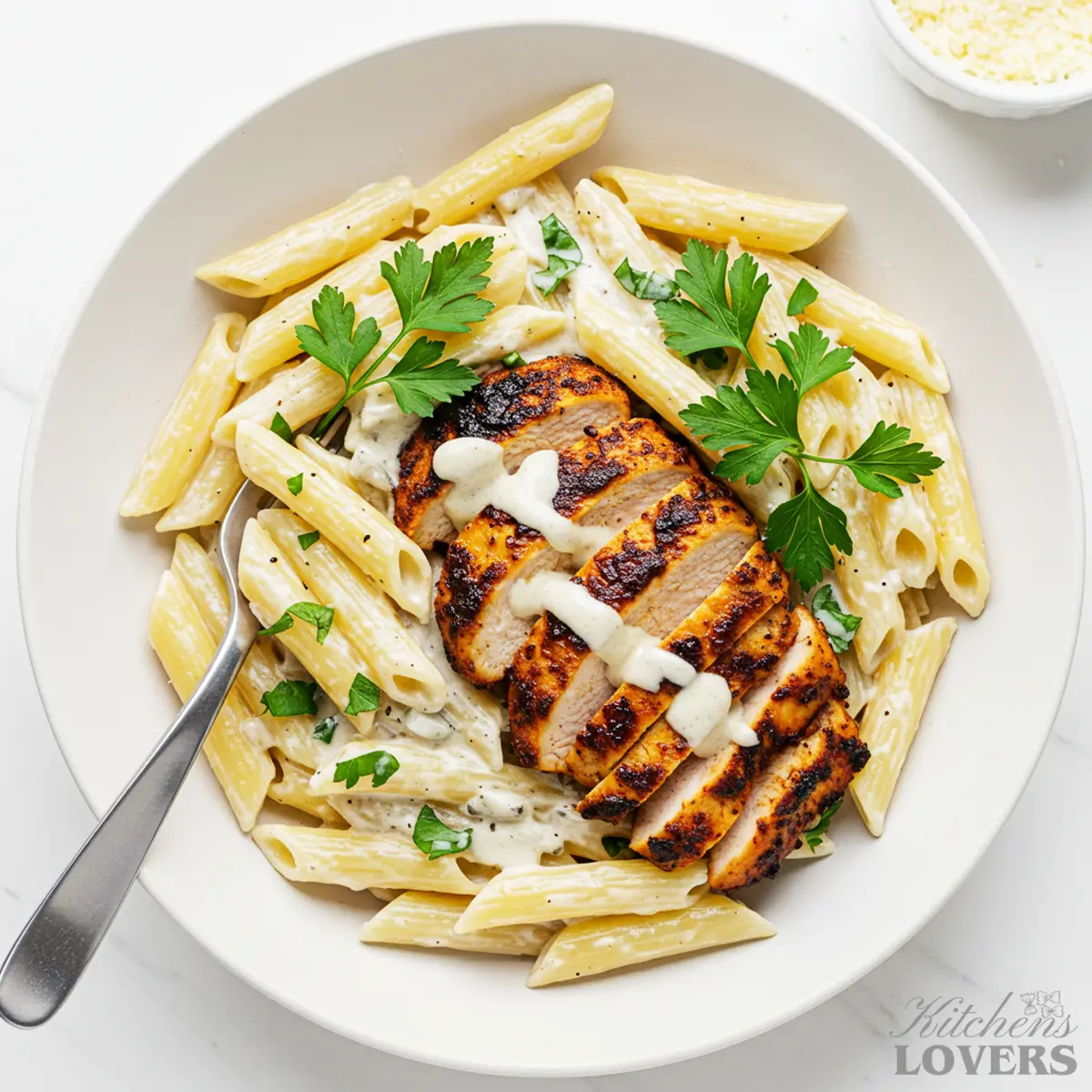 An overhead shot of a white bowl filled with creamy blackened chicken pasta, topped with sliced chicken, fresh parsley, and a side of Parmesan cheese.