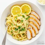 A top-down shot of Creamy Lemon Chicken Pasta in a large white bowl, garnished with lemon wedges, fresh parsley, and slices of seared chicken.
