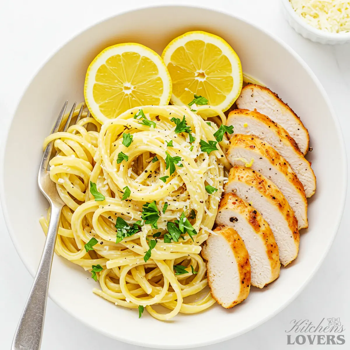 A top-down shot of Creamy Lemon Chicken Pasta in a large white bowl, garnished with lemon wedges, fresh parsley, and slices of seared chicken.