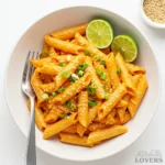 A high-angle overhead shot of a white bowl filled with creamy gochujang rigatoni pasta, garnished with scallions, sesame seeds, and lime wedges.