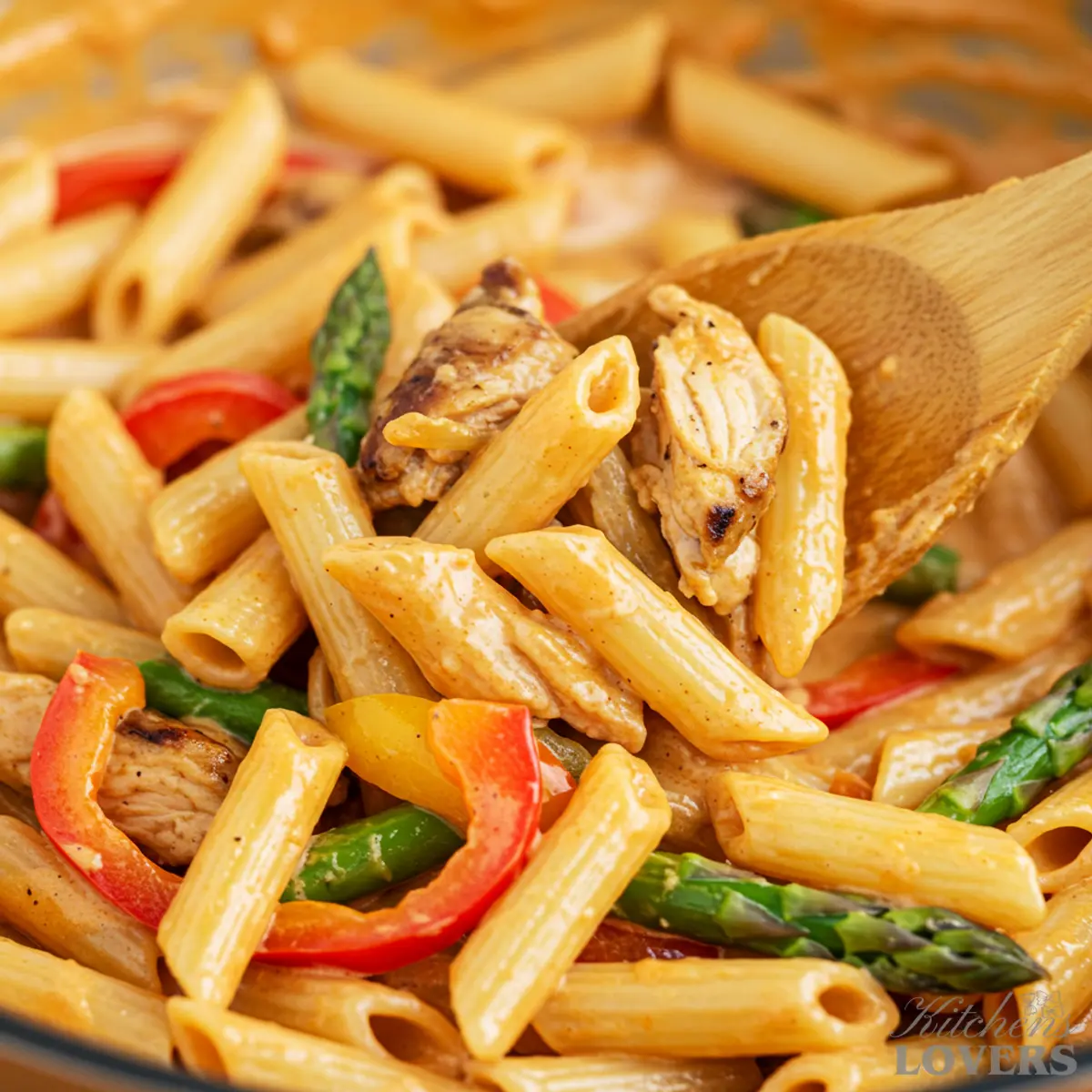 A close-up shot of Jerk Chicken Pasta being mixed with a wooden spoon in a skillet. The creamy orange sauce is visibly coating the penne, chicken, and colorful peppers.