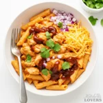 Overhead view of a white bowl of BBQ chicken penne pasta, neatly garnished with a BBQ sauce drizzle, shredded cheddar, diced red onion, and fresh cilantro, on a clean white background.
