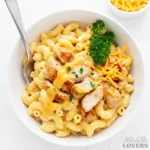 A top-down overview shot of a white bowl filled with creamy chicken mac and cheese, garnished with parsley and paprika, with a fork resting inside.