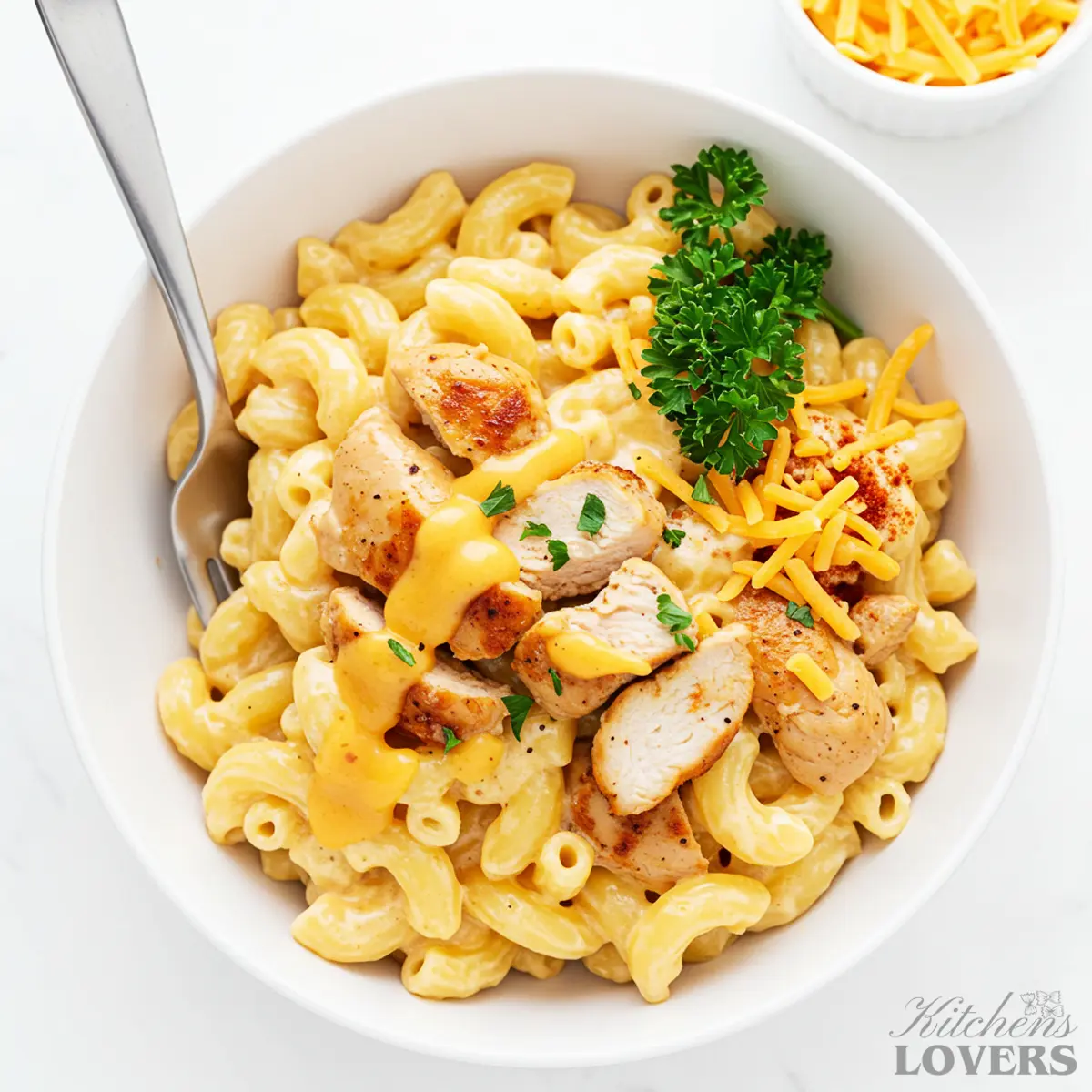 A top-down overview shot of a white bowl filled with creamy chicken mac and cheese, garnished with parsley and paprika, with a fork resting inside.
