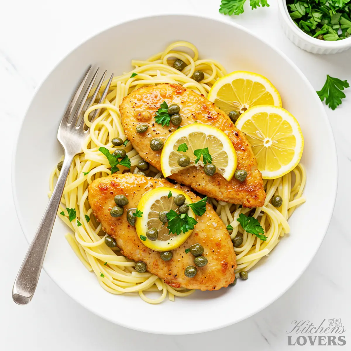 An overhead shot of a white bowl filled with chicken piccata pasta, garnished with lemon wedges, parsley, and capers.