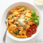 A top-down view of a white bowl filled with creamy sun-dried tomato chicken pasta with rotini, spinach, and Parmesan. A fork rests inside the bowl.