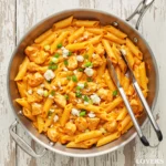 A top-down photo of creamy Buffalo chicken pasta in a skillet, garnished with blue cheese and green onions.
