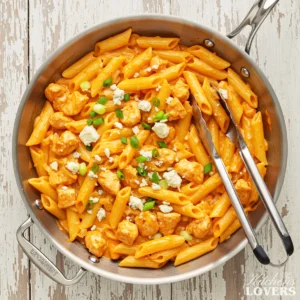 A top-down photo of creamy Buffalo chicken pasta in a skillet, garnished with blue cheese and green onions.